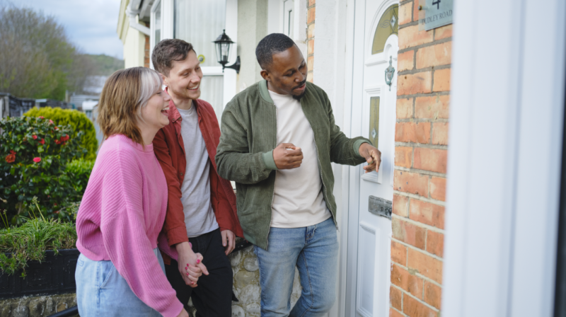 It shows three people looking at a door. Nearest the door is Tayo Oguntonade and beside him is Sam and Bec, a young couple hoping to buy their first home.