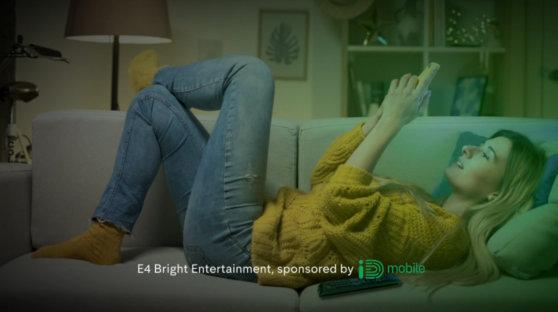 Blonde woman lying on a sofa in a yellow jumper and jeans looking at her phone. On the bottom it says E4 Bright Entertainment, sponsored by iD Mobile (in their green logo).