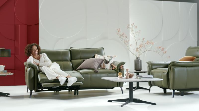 It shows a woman dressed all in white sitting on a olive coloured sofa smiling into the distance. In the room there is also a coffee table, a side table with a plant on it and an armchair. 