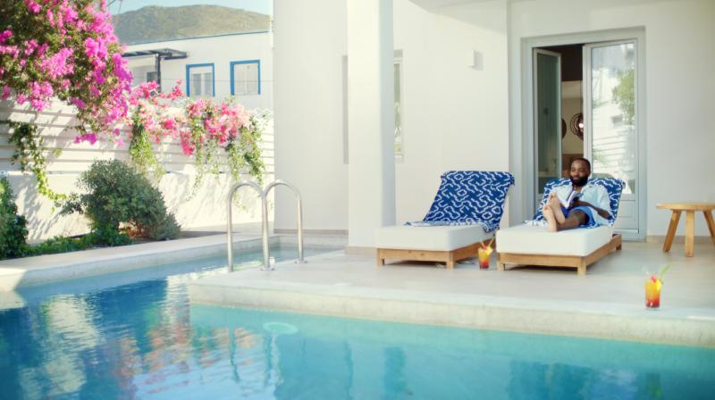 It shows a man reading on a sun lounger by a pool with a white house in the background.