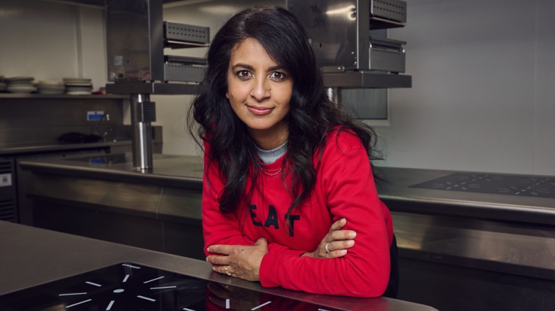 It shows Konnie Huq smiling as she leans on a kitchen counter. She is wearing a red jumper