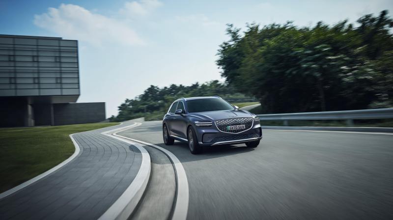 It shows a Genesis car driving on a road bend with trees on one side. 