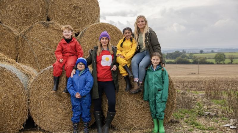 It shows four children in different coloured waterproofs smiling at the camera by hay bales. With them are two adults, Harriet Cowan known for Clarkson’s Farm and comedian Jessica Knapett (The Inbetweeners Movie)