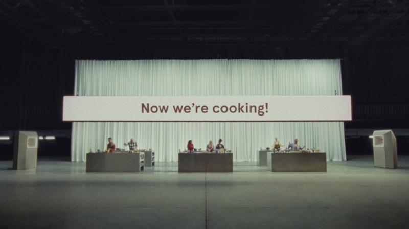 It shows three separate cooking workbenches in a TV studio with people cooking at each of them. Behind them is a large which curtain and on a big screen it says Now We're Cooking!
