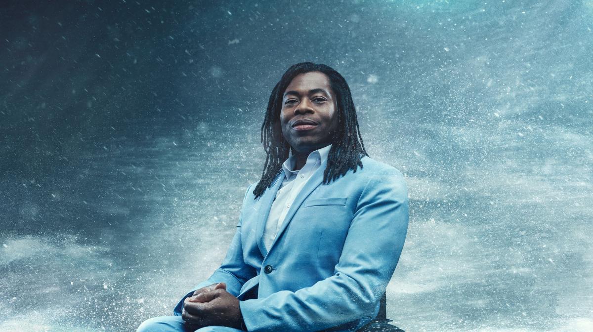 Black male presenter Ade Adepitan wearing a light blue suit sitting in his wheelchair