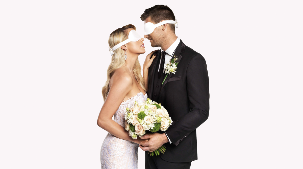 Couple dressed in wedding attire and blind folds leaning in for a kiss.