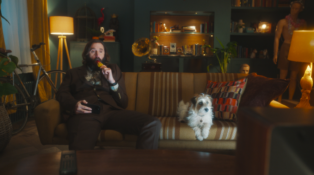 It shows comedian Joe Wilkinson eating a rocket ice lolly on a coach beside a white terrier as they both watch TV. 