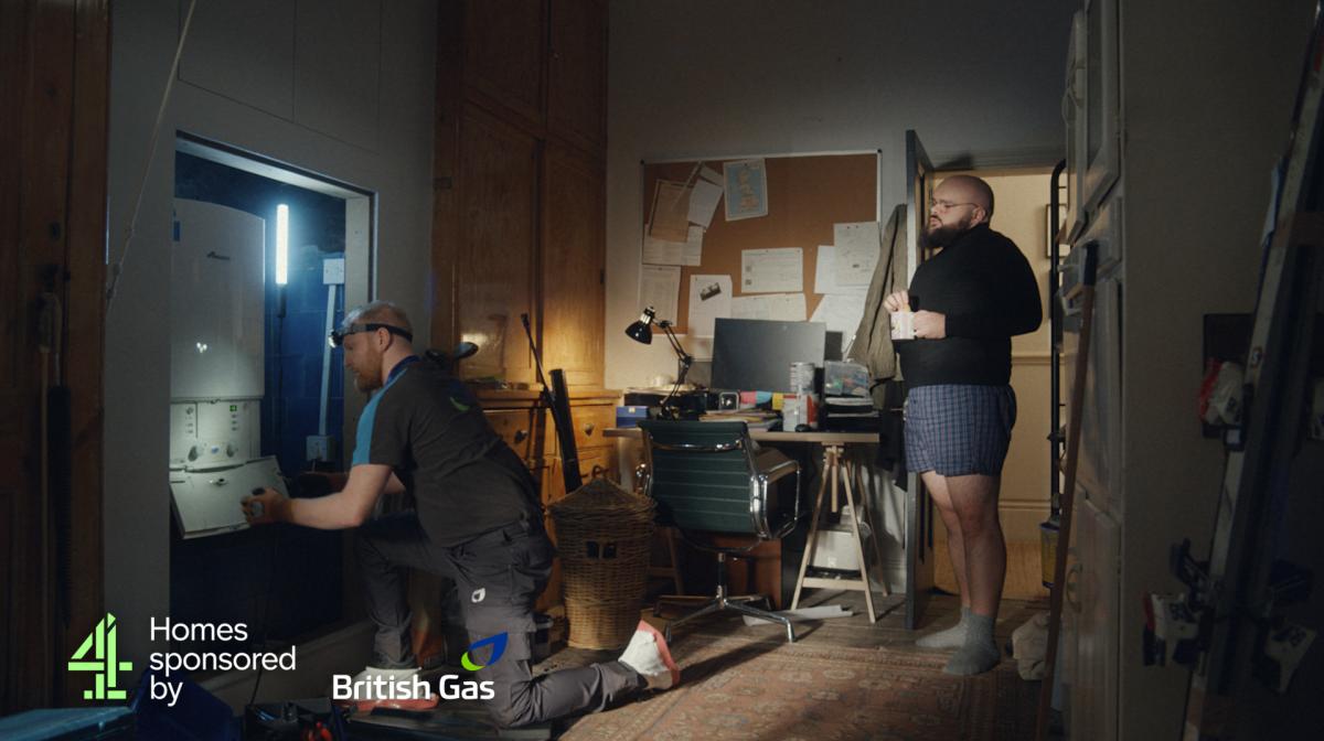 A British gas engineer fixing a boiler as a customer watches on with a cup of tea.