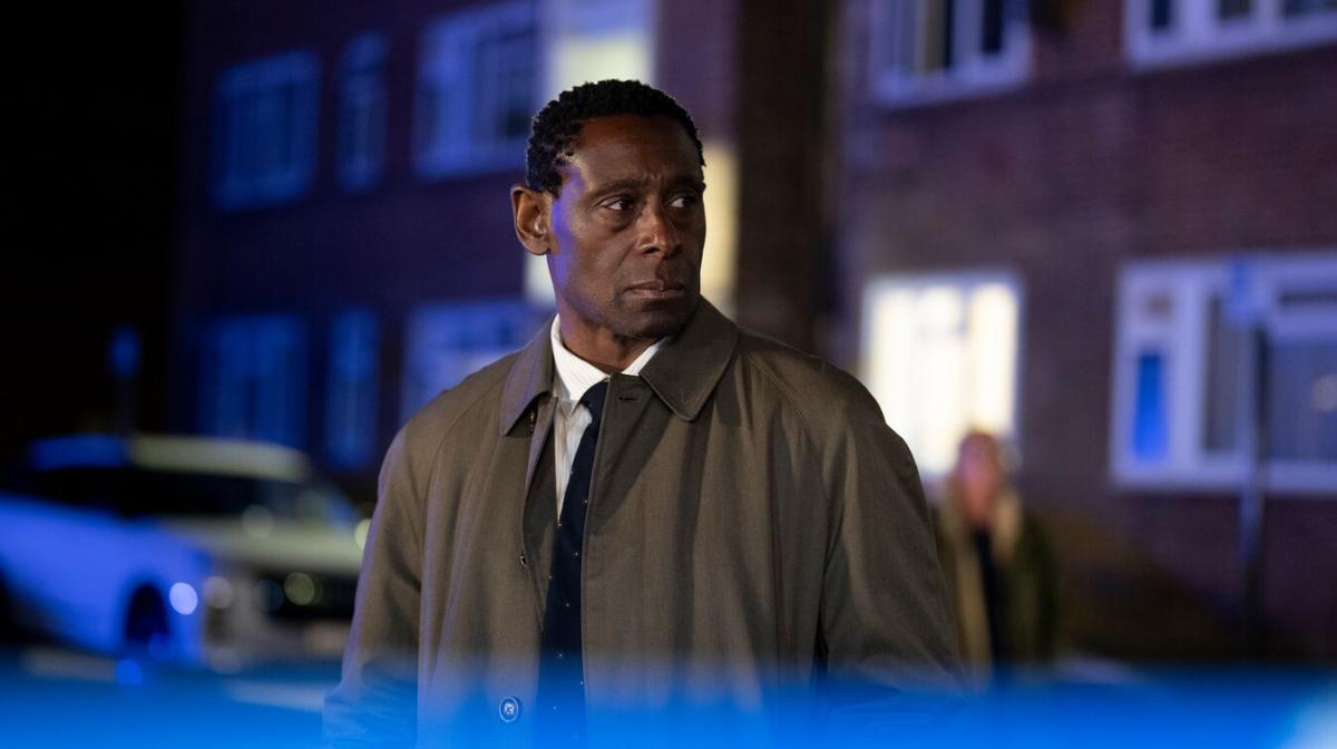 Pierre wearing a coat and stood in a street at night surrounded by a blue light 