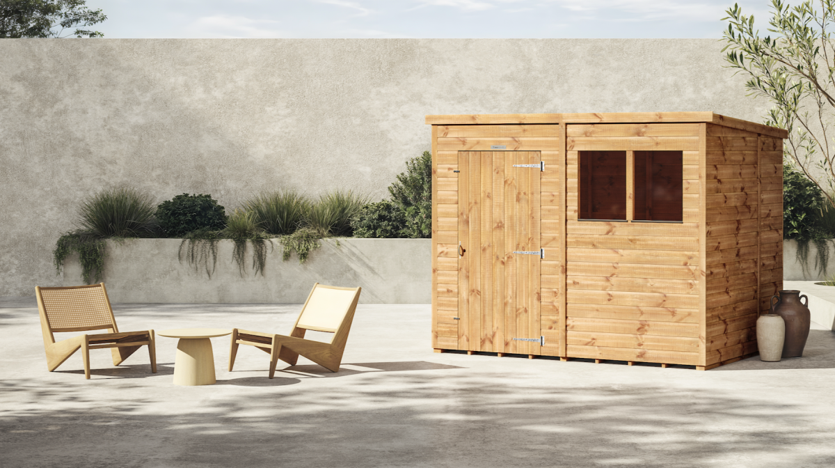 Image shows a polished beige shed in a stone garden setting with a beige table and 2 rattan chairs on the left hand side. Potted plants are seen in the background and next to the shed. The sun is shining on the sheds.