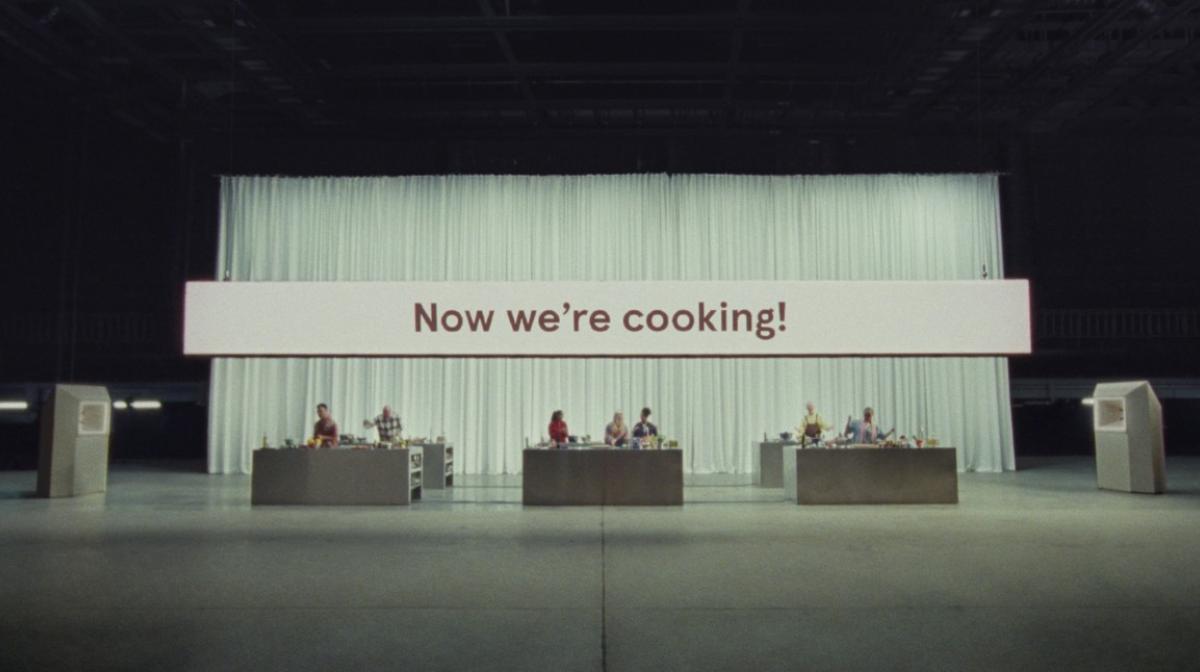 It shows three separate cooking workbenches in a TV studio with people cooking at each of them. Behind them is a large which curtain and on a big screen it says Now We're Cooking!