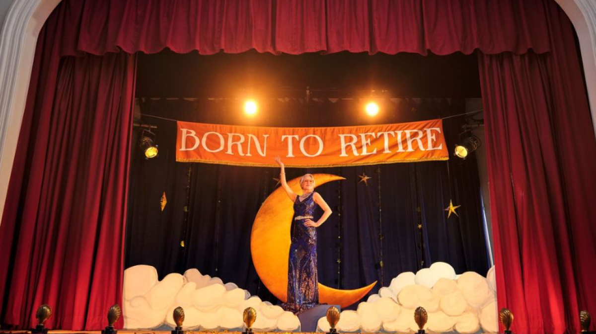 Image shows a female in a sparkly blue dress on a theatre like stage production. There are red curtains pulled to the side. Behind her is a crescent moon and clouds with a banner noting the phrase 'Born to Retire' above her.