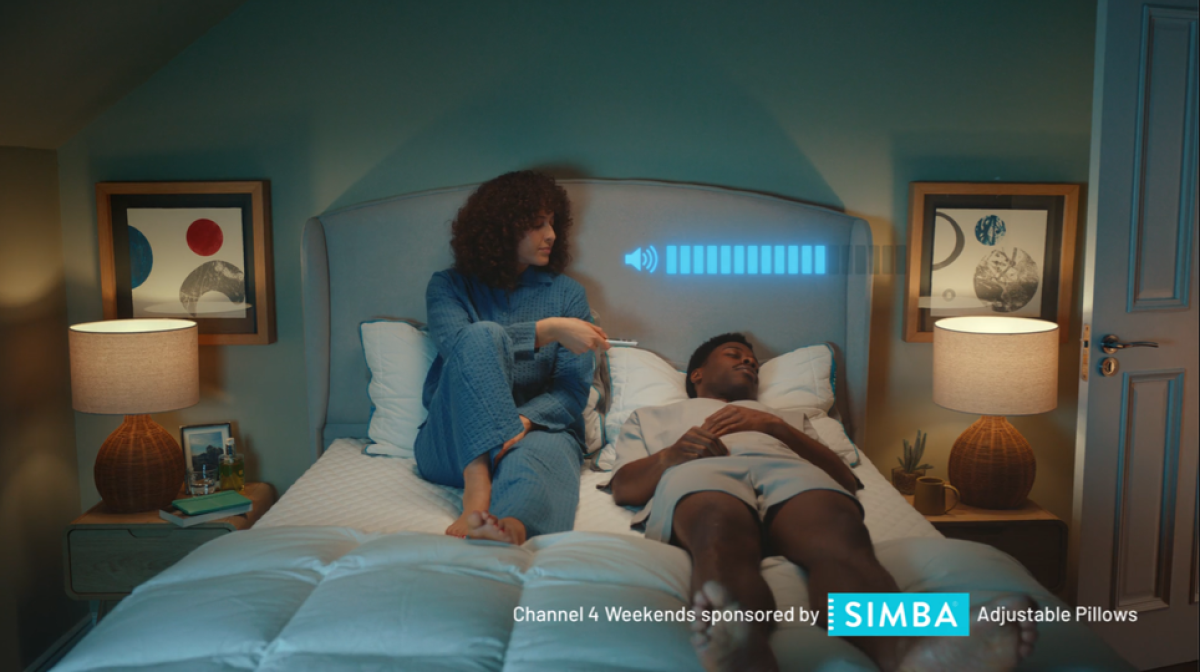 A couple are on a Simba mattress in a bedroom. The male on the right hand side is lying down sleeping and the female on the right hand side is putting the volume down, shown by a volume graphic, of the television she is watching.