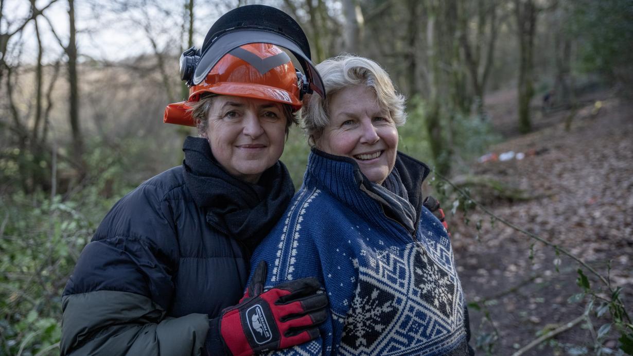 In a close up shot, Sandi Toksvig and her wife Debbie stand in the middle of a woodlands. Debbie is on the left embracing Sandi on the right. Sandi has short blonde hair, and wears a blue and white Fair Isle quarter-zip sweater. Debbie wears a tree surgeon's hat, ear defenders and gloves with a black puffer coat. Both women are smiling directly to camera. 