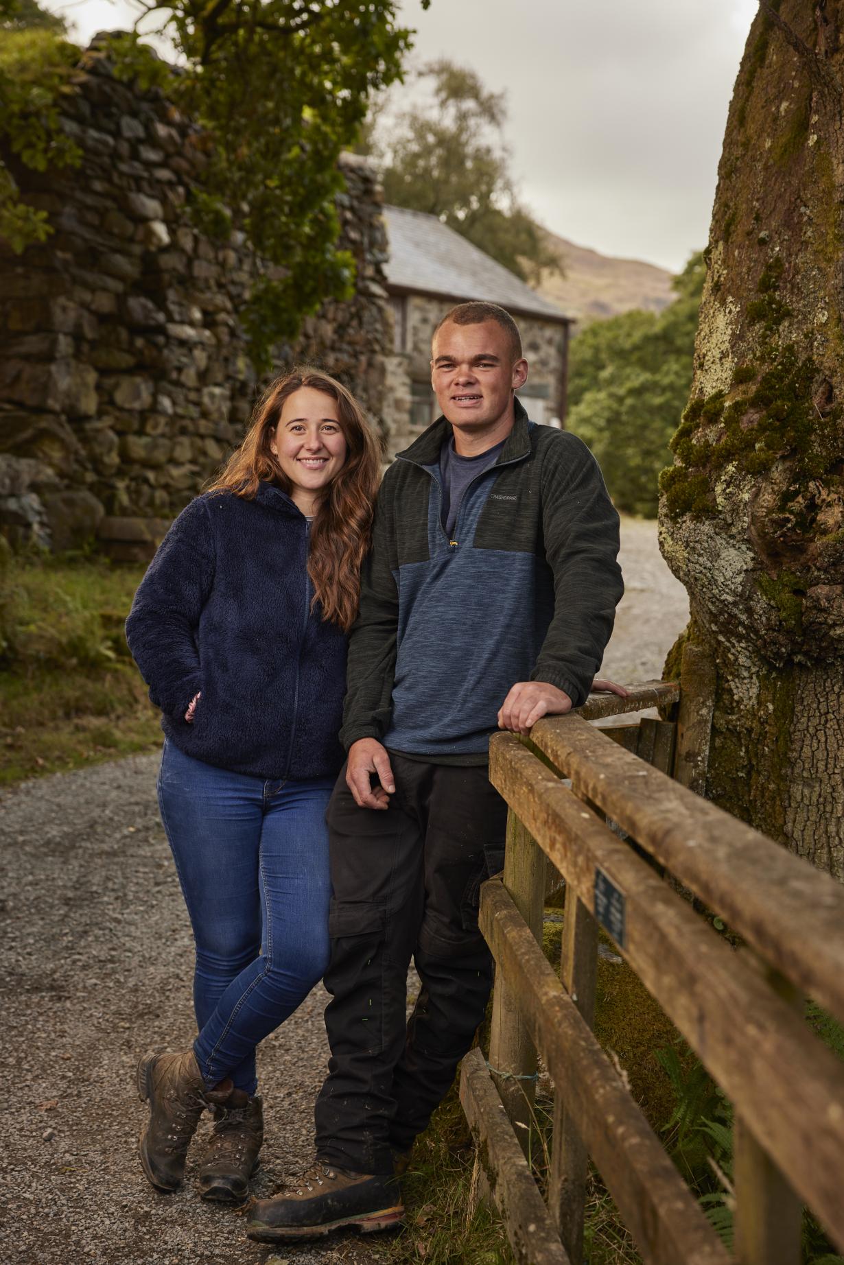 Winners Sara on the left and Ioan on the right, lean against a wooden fence - captured full length smiling at the camera. Pictured on the road leading up to a farmhouse, Sara wears a blue fleece and jeans, and Ioan grey fleece and black worker trousers.