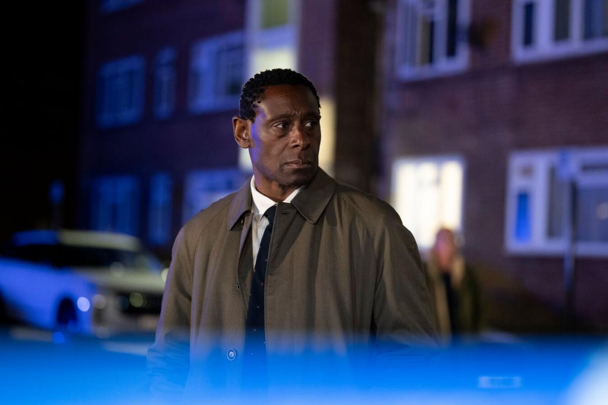 Pierre wearing a coat and stood in a street at night surrounded by a blue light 