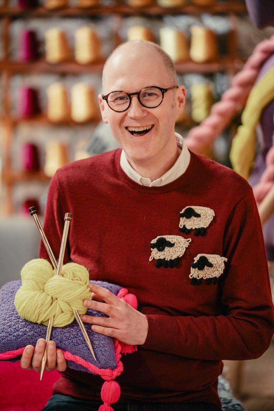 Game of Wool winner Holger wears a red jumper with three sheep badges attached and holds a cushion with a ball of wool and a pair of gold knitting needles on it.