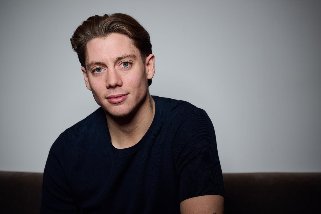 Sam wears a dark short-sleeve shirt sits indoors against a plain wall. The lighting is subdued, casting soft shadows, and the focus is on the upper body and posture. 