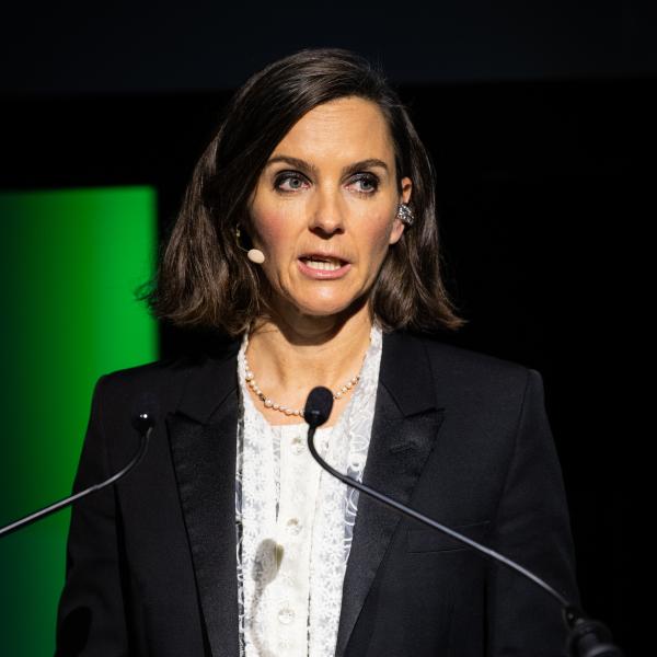 It shows Alex Mahon speaking into a microphone and standing behind a lectern. 