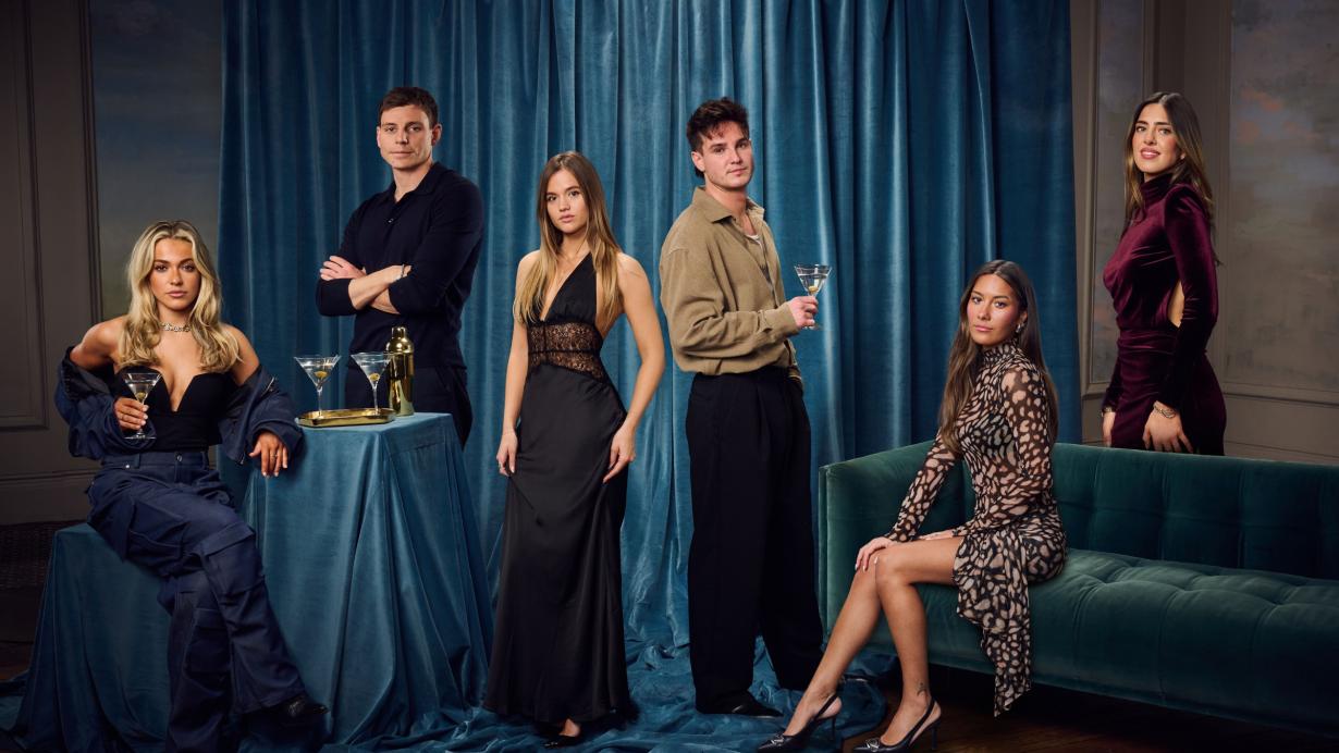 (L-R) Jazz, Tristan, Jules, Sam Prince, Inga and Yas pose in an indoor setting with blue curtains as a backdrop. All in evening attire, the cast wear a range of black and burgundy dresses, tailored shirts, and trousers. Two people hold martini glasses, and a small table draped in blue fabric displays additional glasses and a cocktail shaker. One woman sits on a green velvet sofa, another leans against the covered table, while the others stand confidently, facing the camera. 